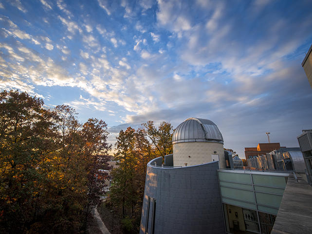 A wide angle shot of a cloudy sky with the fall leaves and a Mason campus building in the forefront.