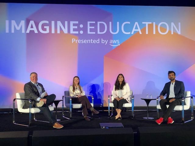 Maya Chatterjee sitting in a row with other AWS Imagine EDU student speakers in front of a colorful screen backdrop.