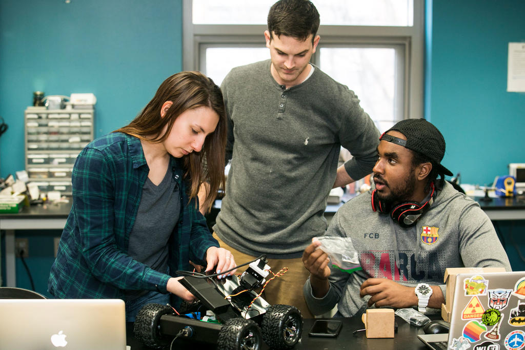 One female student and two male students in a college robotics lab build a mobile robot and try to connect the electrical wires.