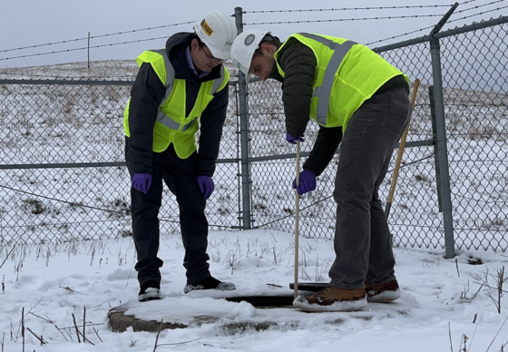 Two people in a snowy landscape drop a measuring device into a sewer to test for continents. 