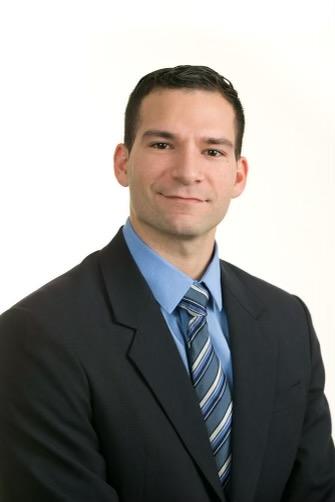 Bryan Vega poses for the camera in a black suit and blue shirt and tie. 