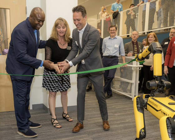 Three people are cutting a ribbon, one side of which is held by a robotic quadruped