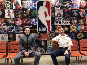 An army cadet and officer sit in front of a wall with a variety of NBA posters